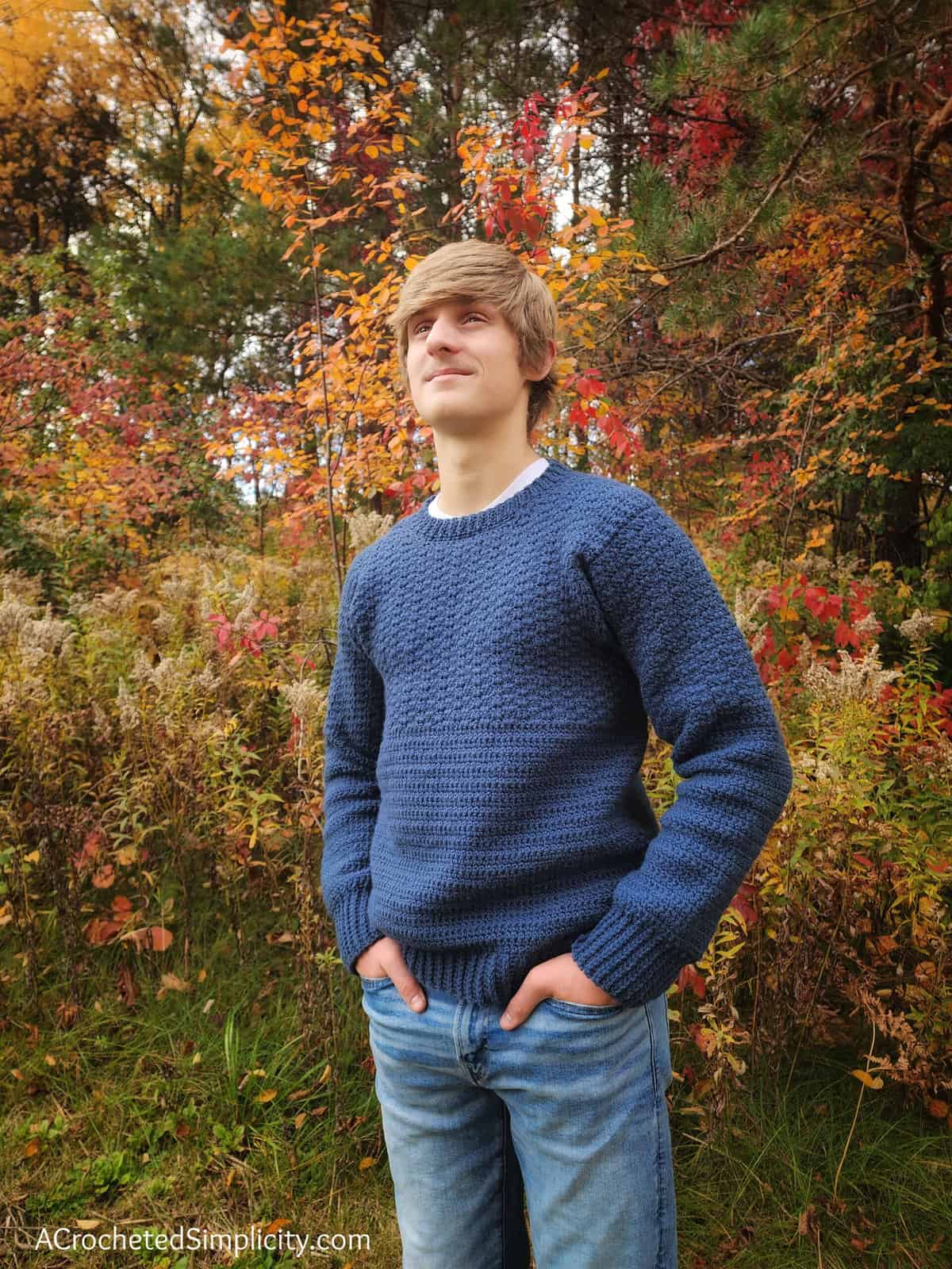 Young man modeling a blue crew neck crochet pullover with fall leaves in background.