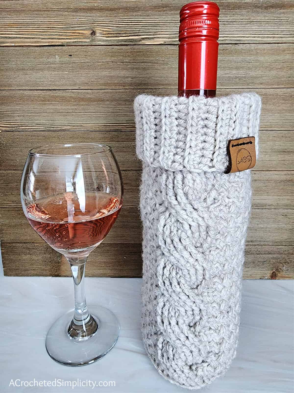 Oatmeal colored cabled wine cozy with wine inside sitting next to a wine glass with wine.