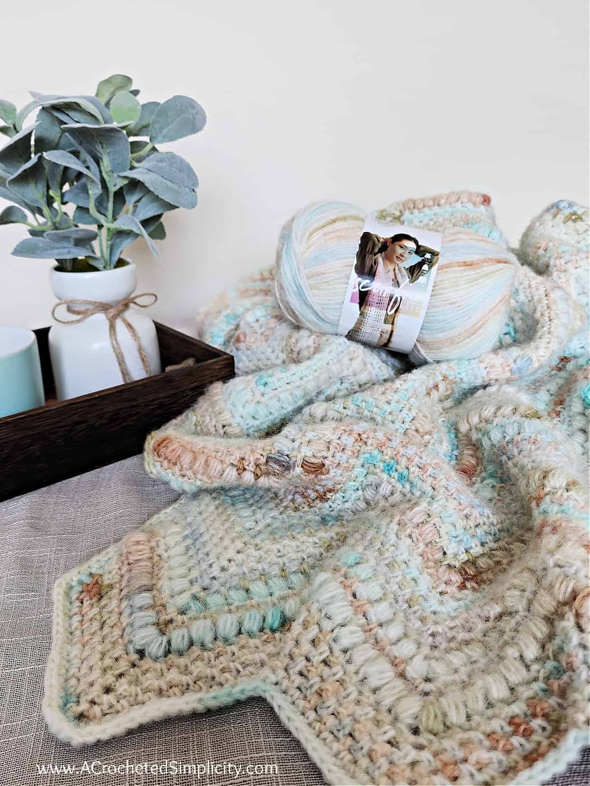 Ripple crochet blanket in beachy colors laying with ball of yarn and lamb's ear plant.