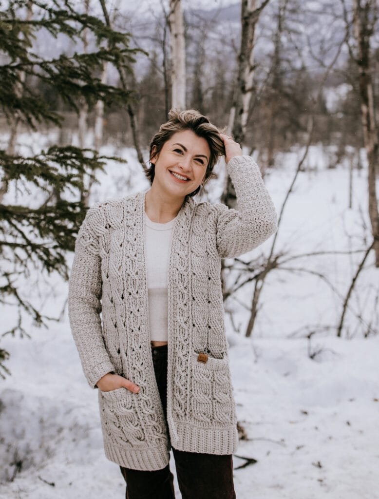 Woman wearing light taupe cabled cardigan next to pine tree.