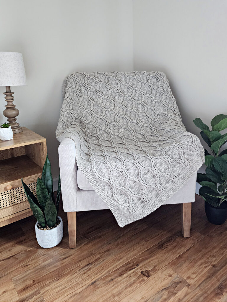 Light grey crochet cable blanket draped over a small sitting chair with green plants nearby.