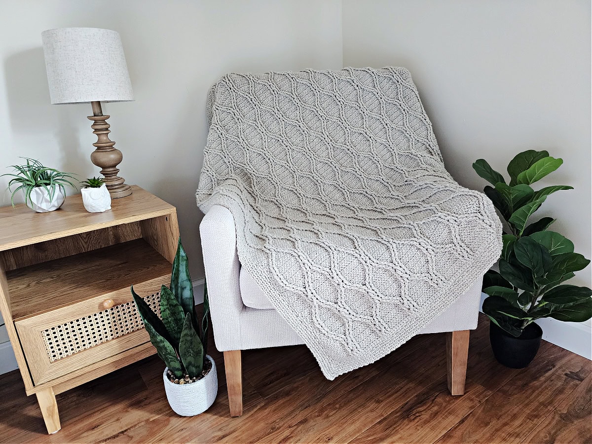 Crochet cable throw draped across a small armchair next to a small wooden table.