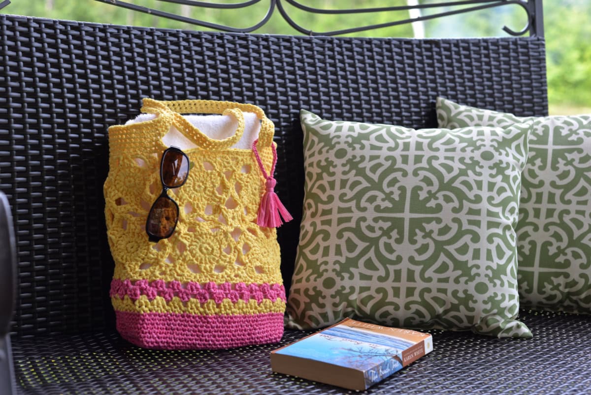 Pink and yellow flower motif crochet tote bag sitting on a porch swing with a book and green pillows.