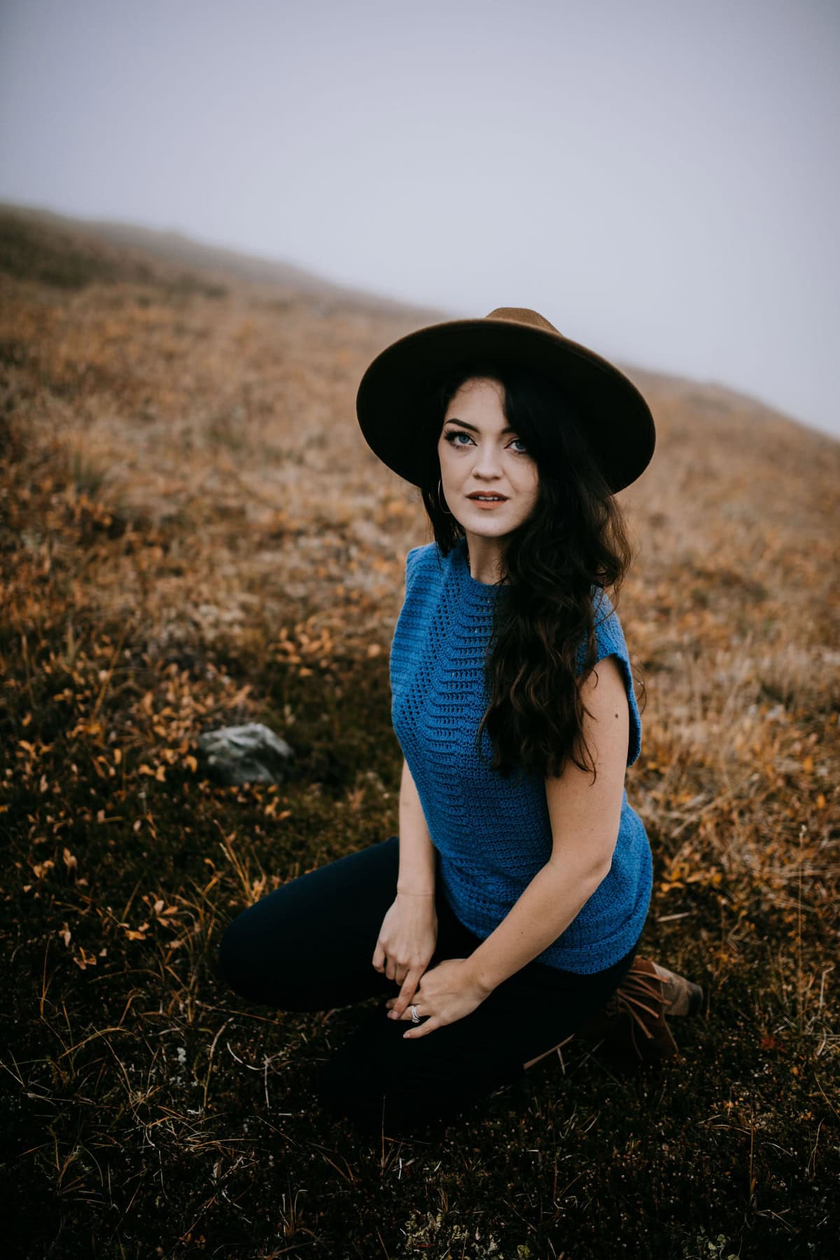 Young woman wearing a hat and teal summer crochet top and kneeling on a mountain.