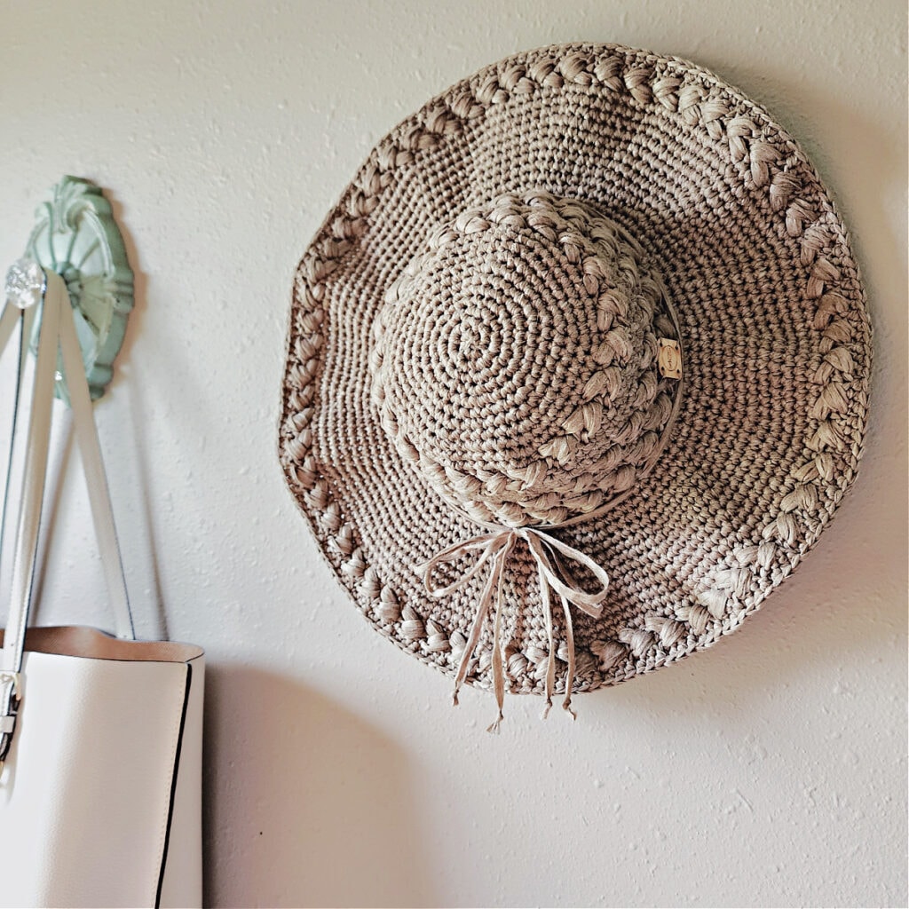 A beige crochet sun hat with a wide brim hanging on a hook on the wall.