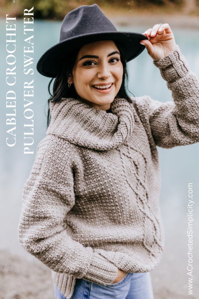 Close up of a crochet cable sweater pullover with a cowl neck modeled by a woman in a black hat.