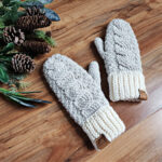 Pair of neutral color cable crochet mittens with leather tags laying next to pine cones and leaves.