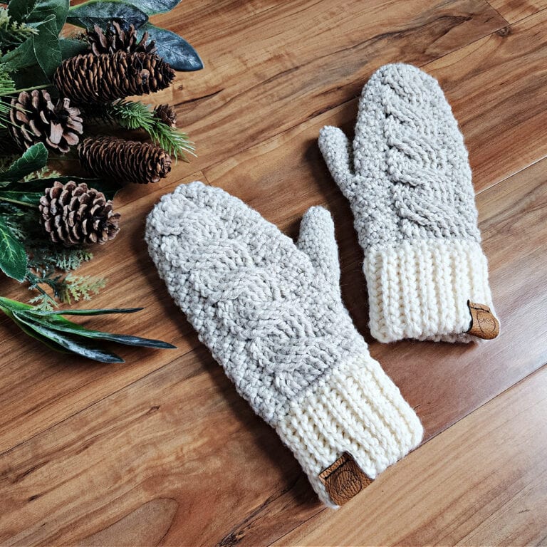 Pair of neutral color cable crochet mittens with leather tags laying next to pine cones and leaves.