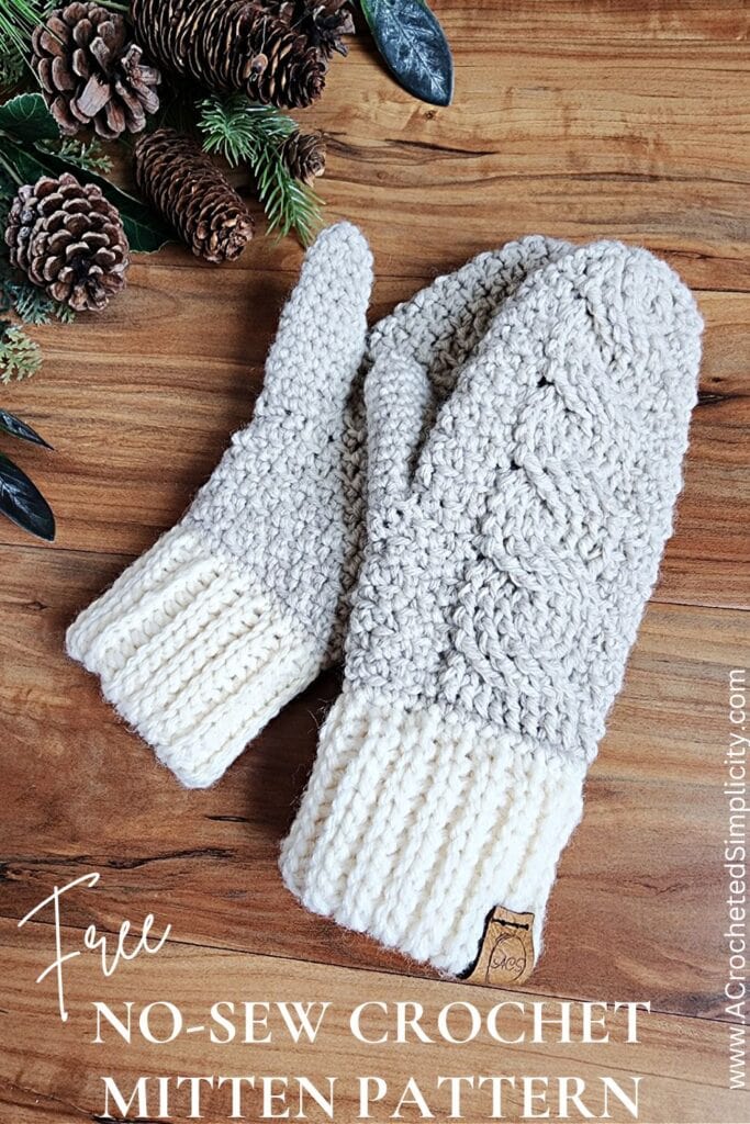 Pair of cable crochet mittens laying on top of one another next to pine cones on wood surface.