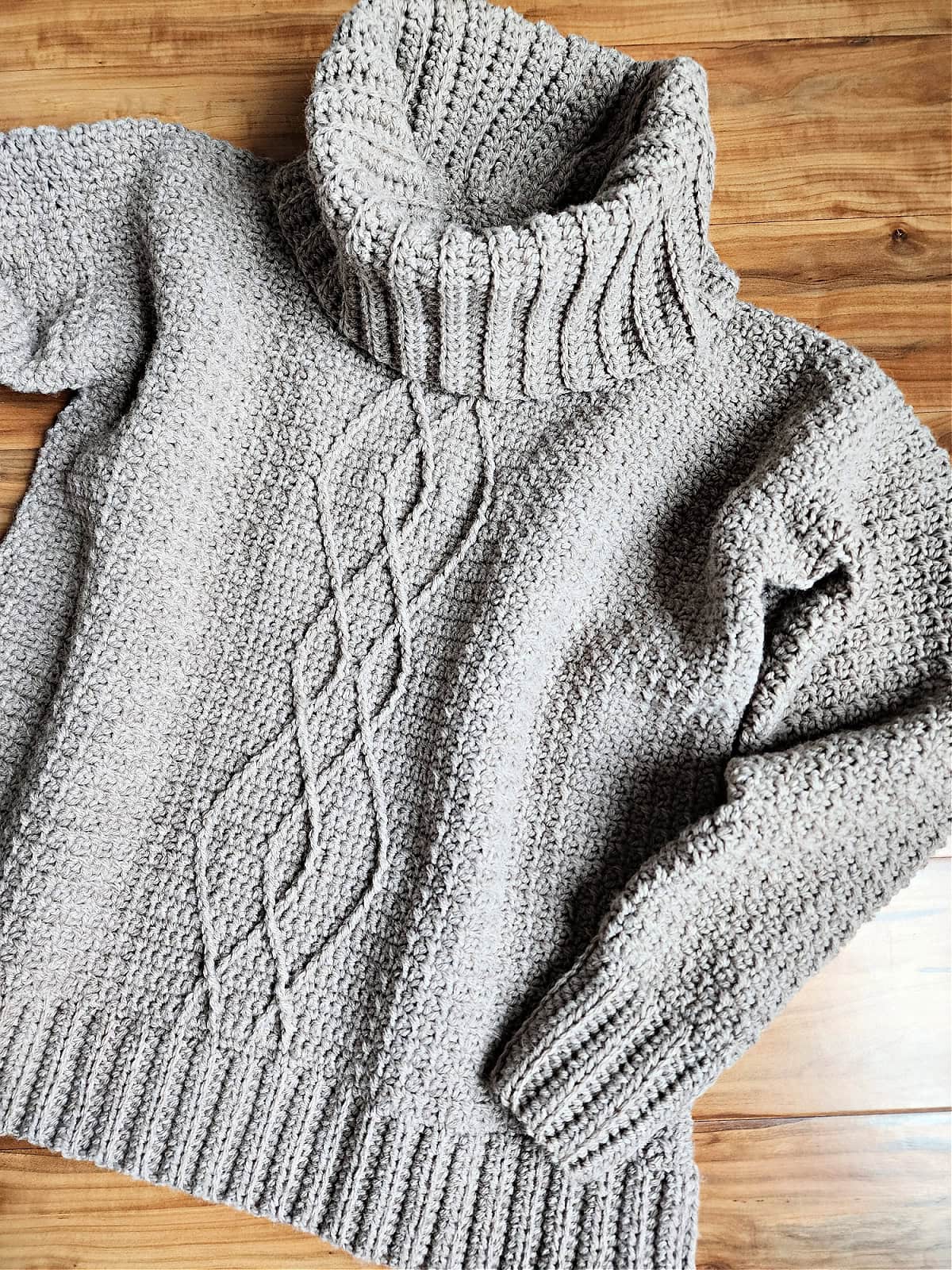 A cable crochet sweater pullover with a cowl neck laying on a wood surface.