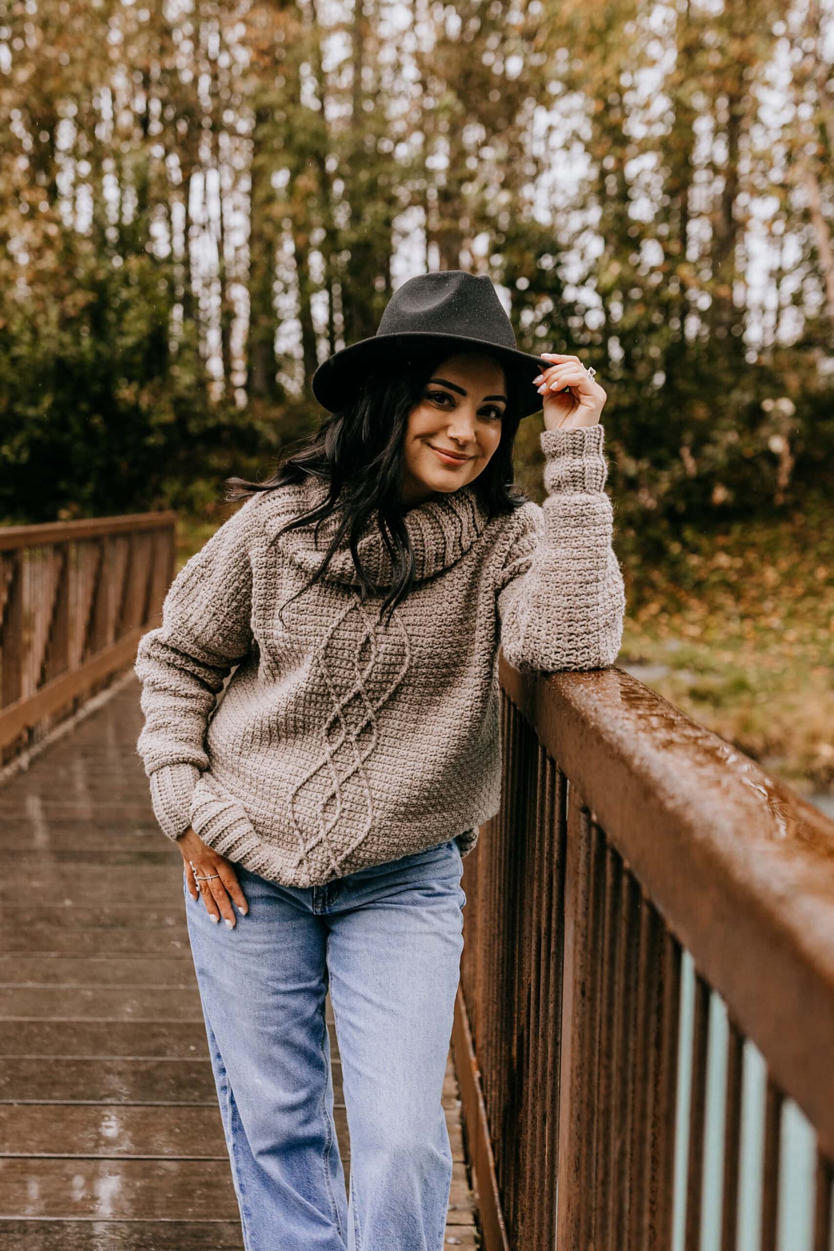 Woman standing on a wooden bridge modeling a crochet cable sweater with a loose cowl neck.