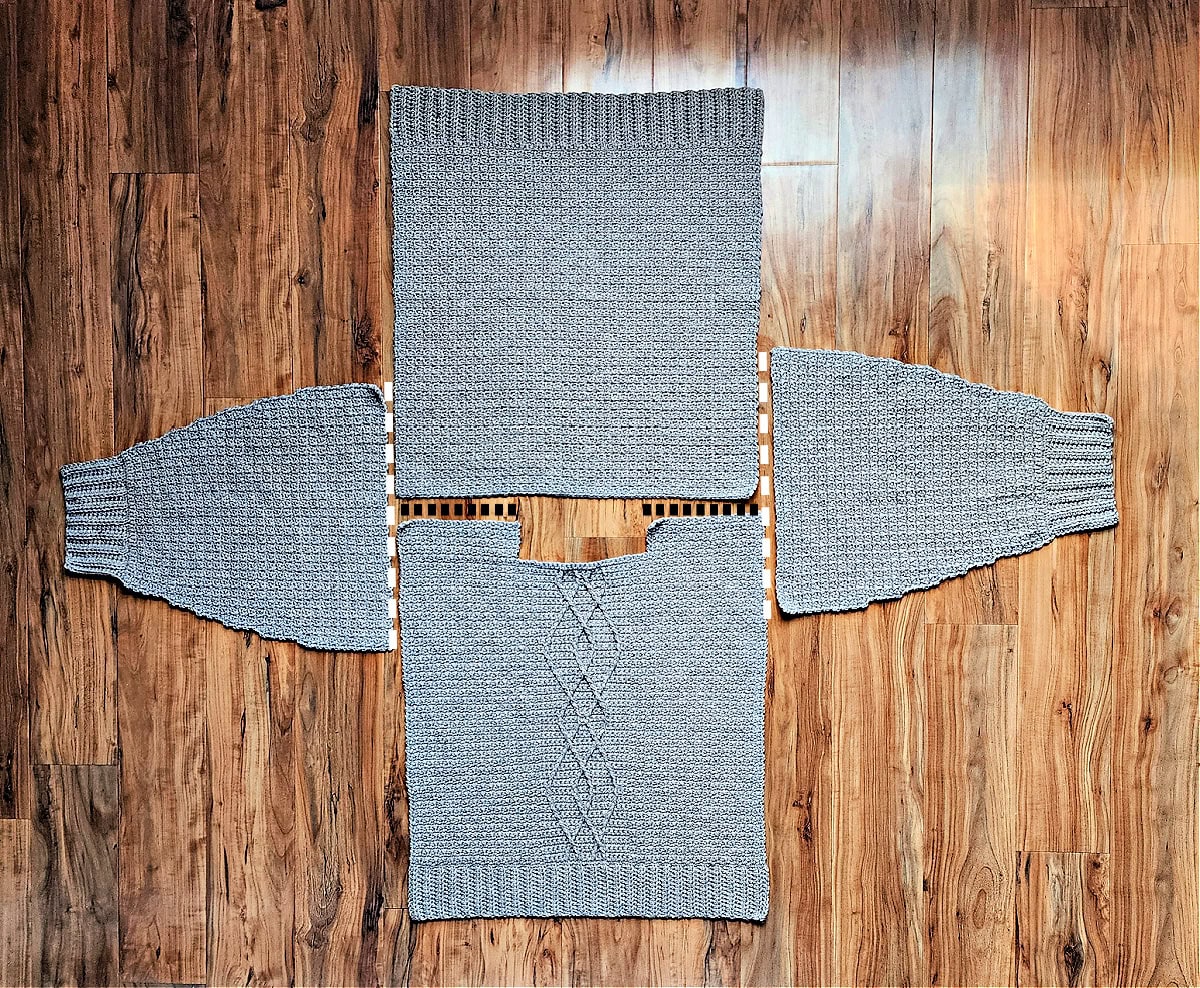 Four panels of a cable crochet sweater laid flat on the floor with dotted lines to show where to seam.