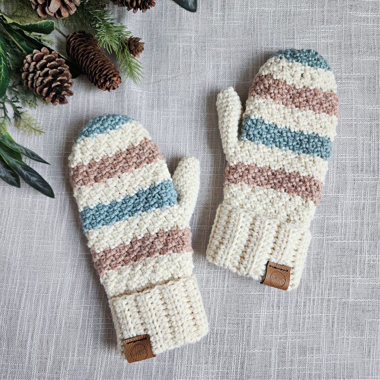 A pair of striped crochet mittens laying on a cloth with pine cones.