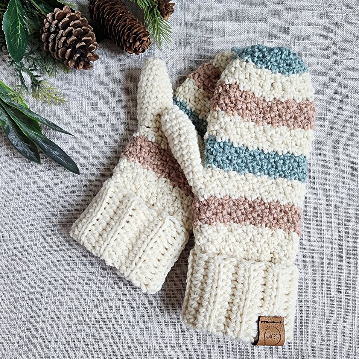 One striped crochet mitten laying on top of another one next to pinecones.