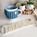 Knit-look crochet mug cozy wrapped around a coffee cup sitting on the counter next to a mug cozy laid flat.