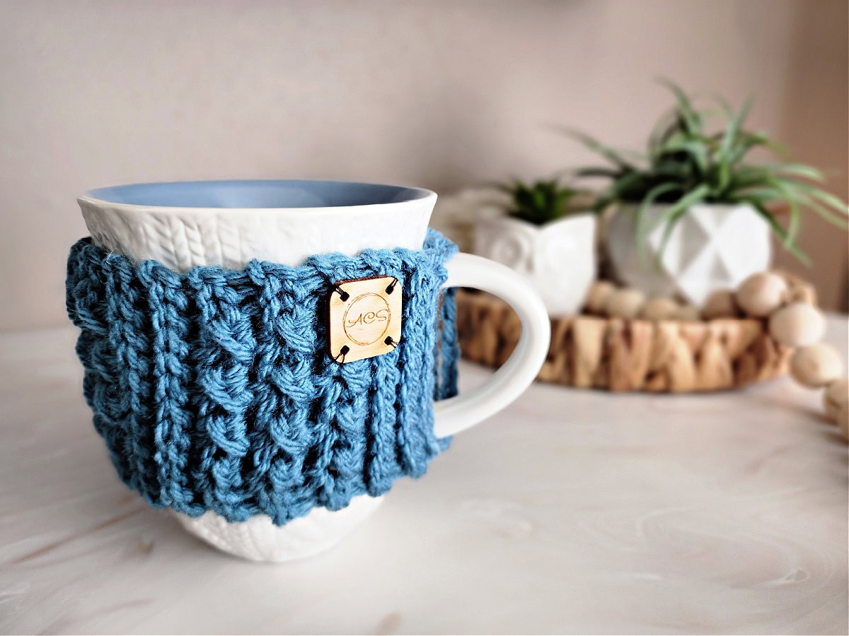 A crocheted coffee mug cozy with a wooden tag wrapped around a white cabled coffee mug.