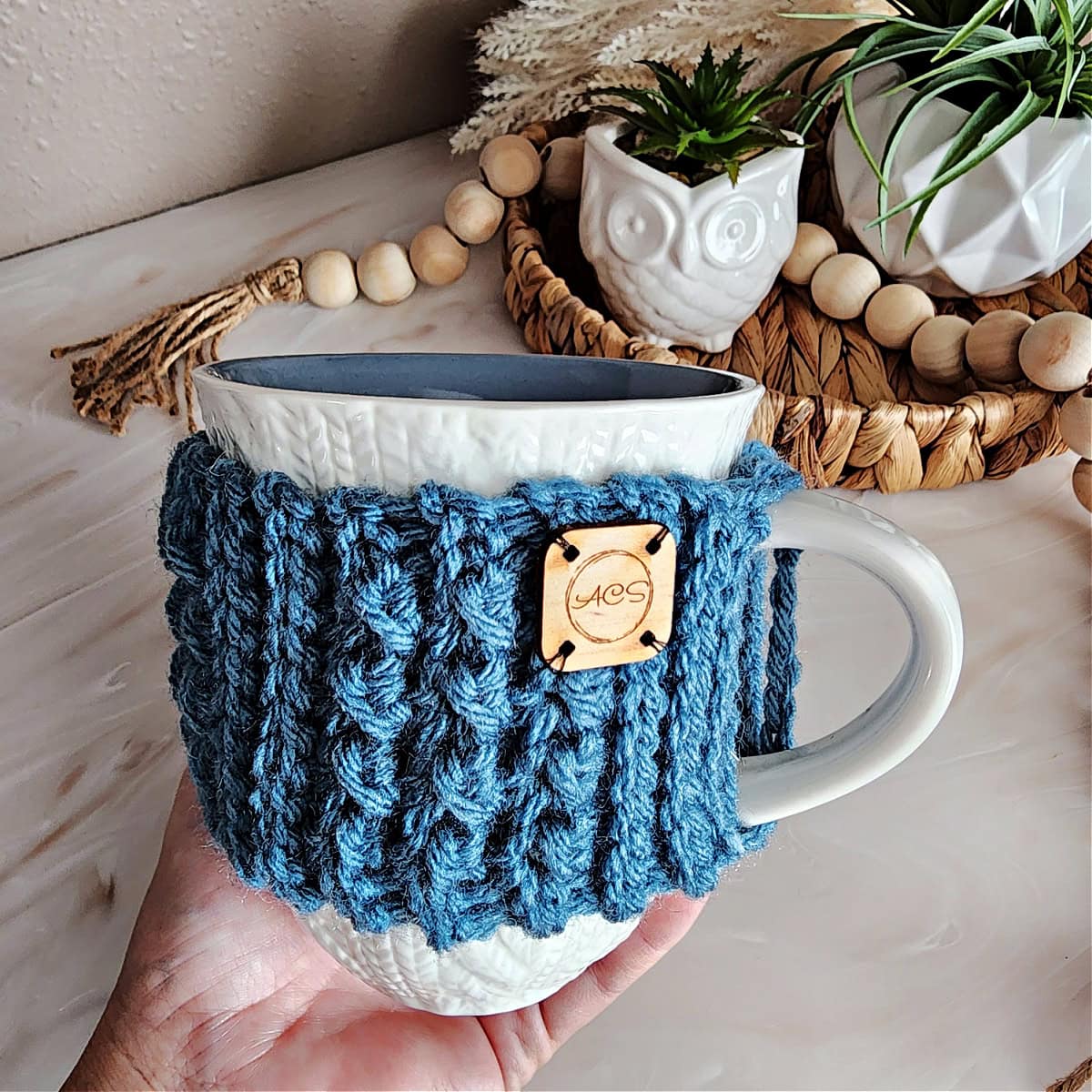 A woman's hand holding a coffee mug with a blue crocheted mug cozy wrapped around it.