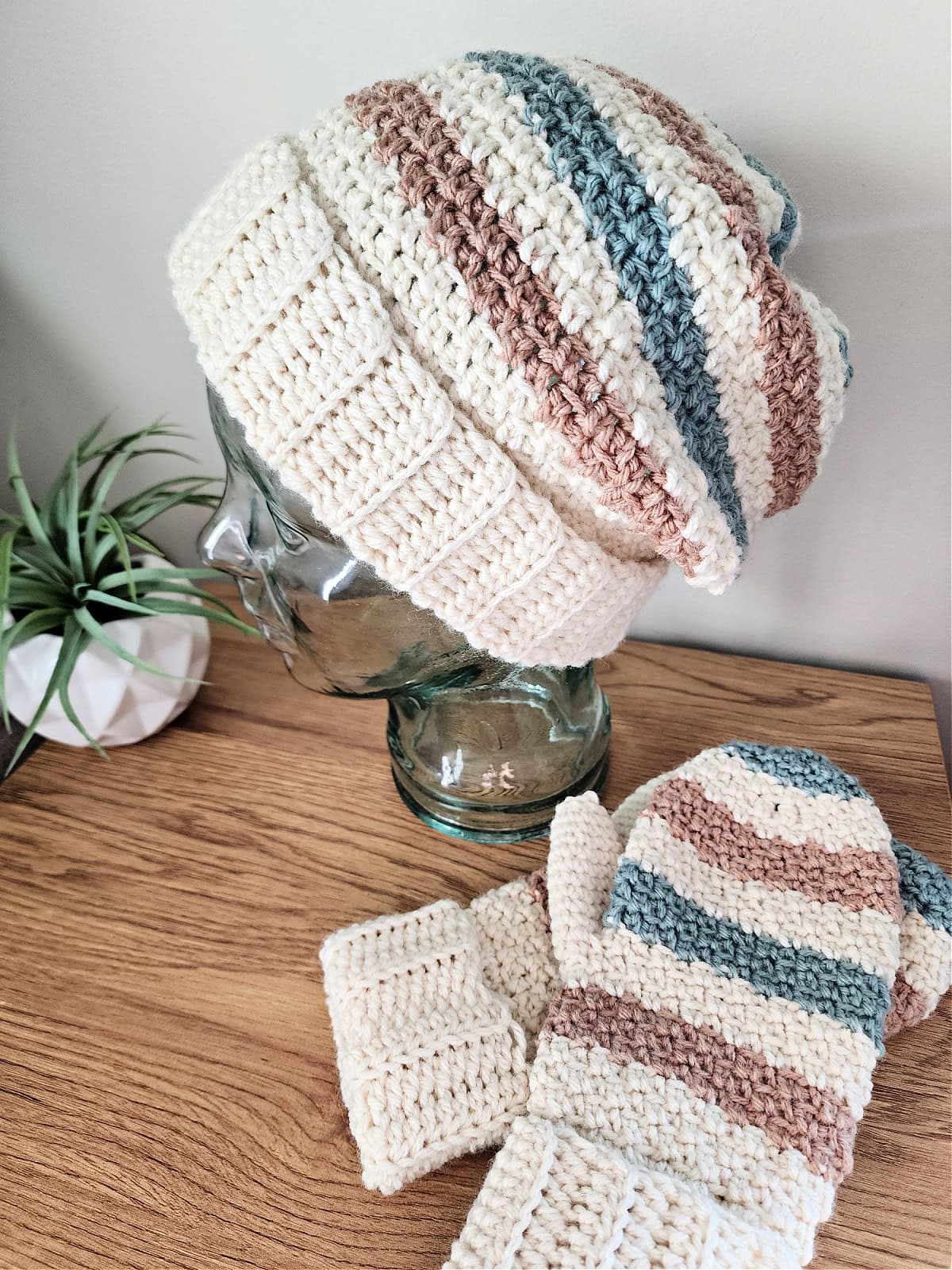 A cream and stripe slouchy beanie crochet pattern on a glass mannequin with striped crochet mittens laying next to it.