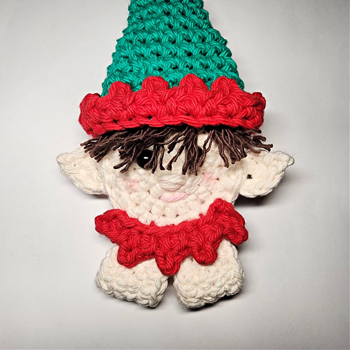 A green and red elf hat with brown hair being attached to the top of the crocheted elf.