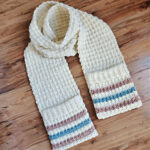 A cream colored crochet pocket scarf laying on a wood surface.