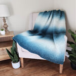 Gradient crochet blanket draped across a white chair with plants next to it.