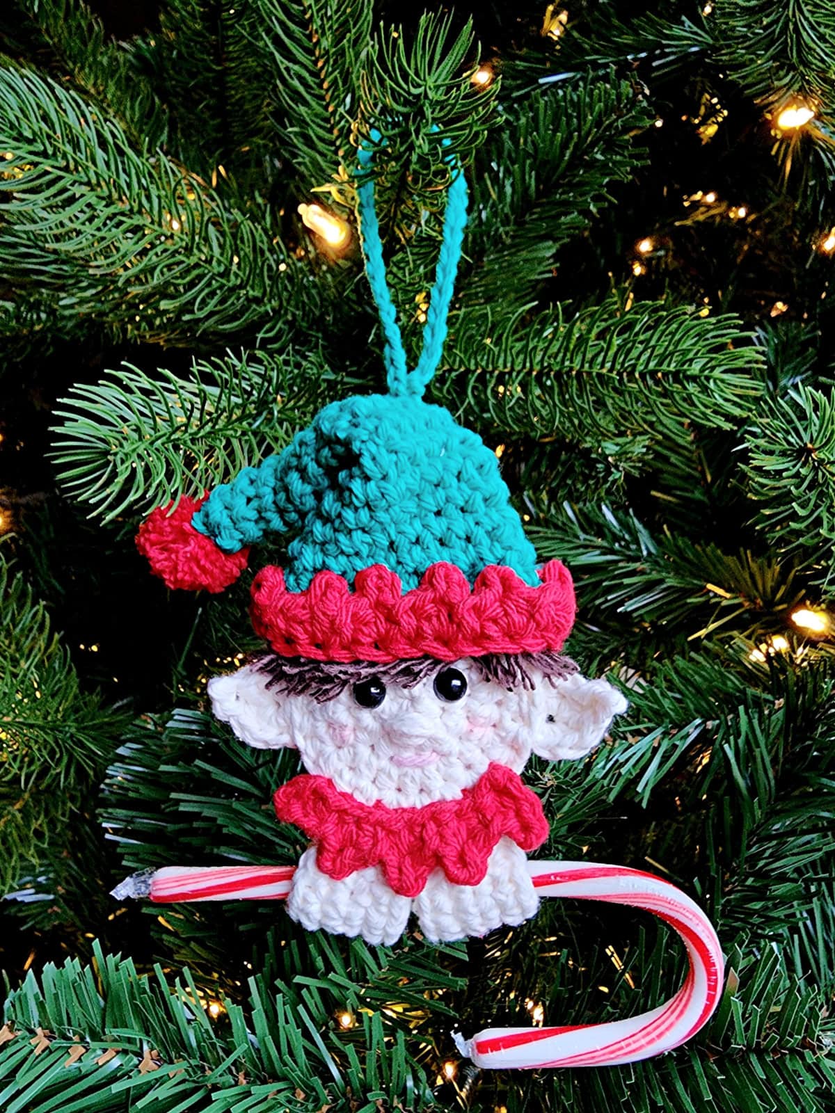 Crochet elf ornament holding a candy cane and hanging on a Christmas tree.