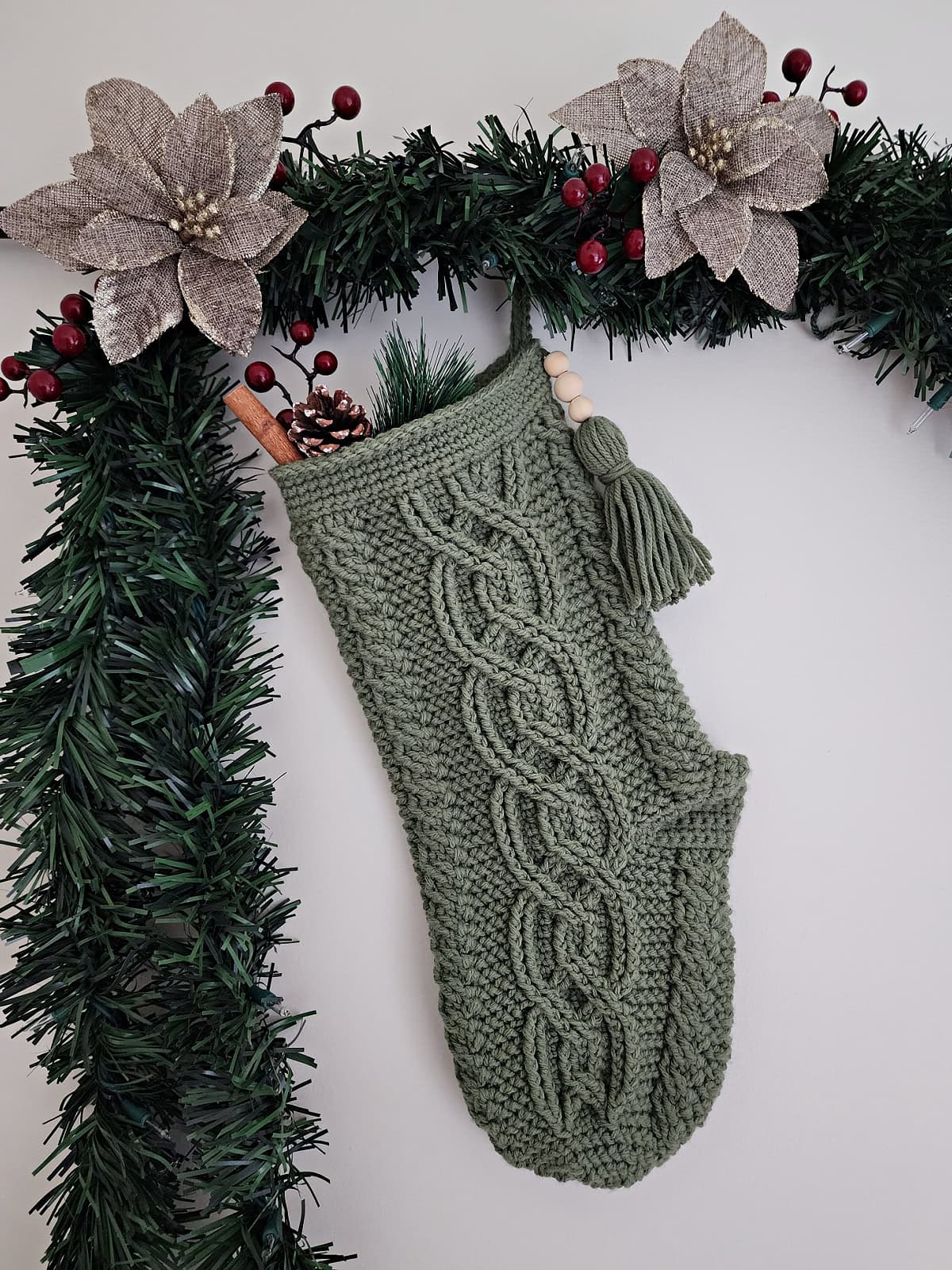 Green cabled crochet stocking with celtic cables hanging up with garland and poinsettias.