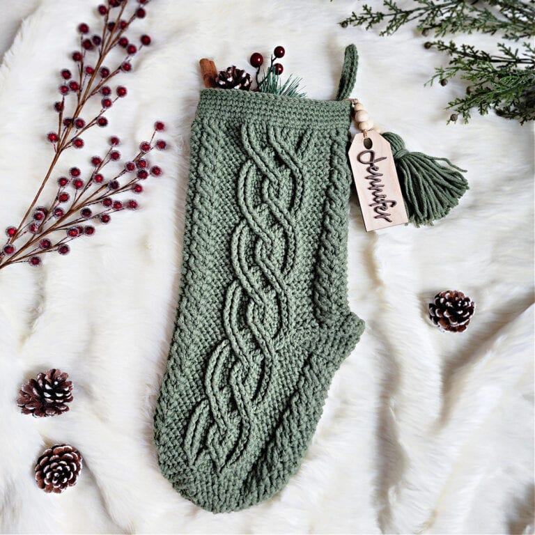 An olive green crochet Christmas stocking with celtic cables laying on a cream fur blanket with berries and pine cones.