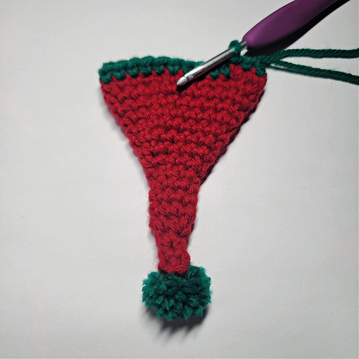 A purple crochet hook adding a green border to the red crochet elf hat.