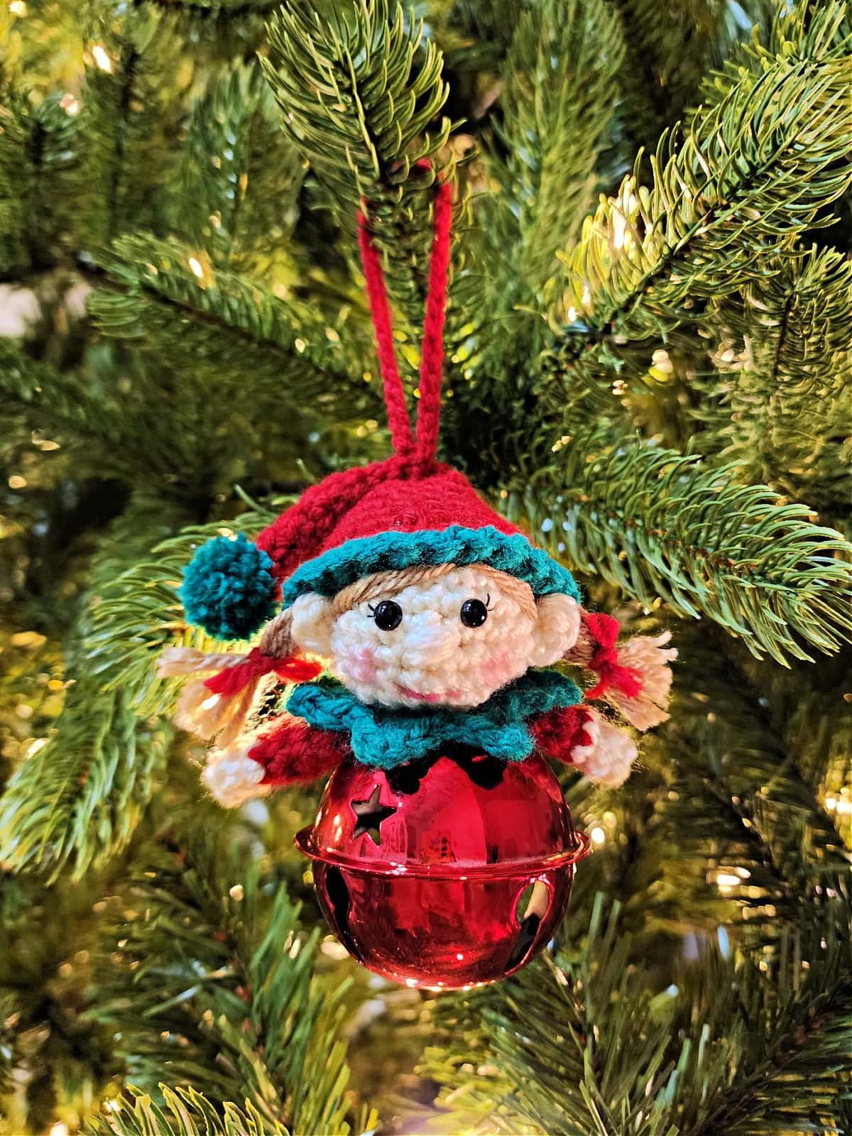 A sweet little crochet girl elf jingle bell ornament hanging on a Christmas tree.