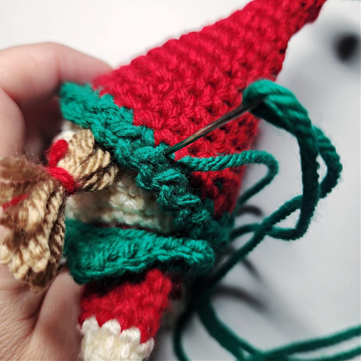 A yarn needle with green yarn showing how to attach a hat to the crochet elf ornament.