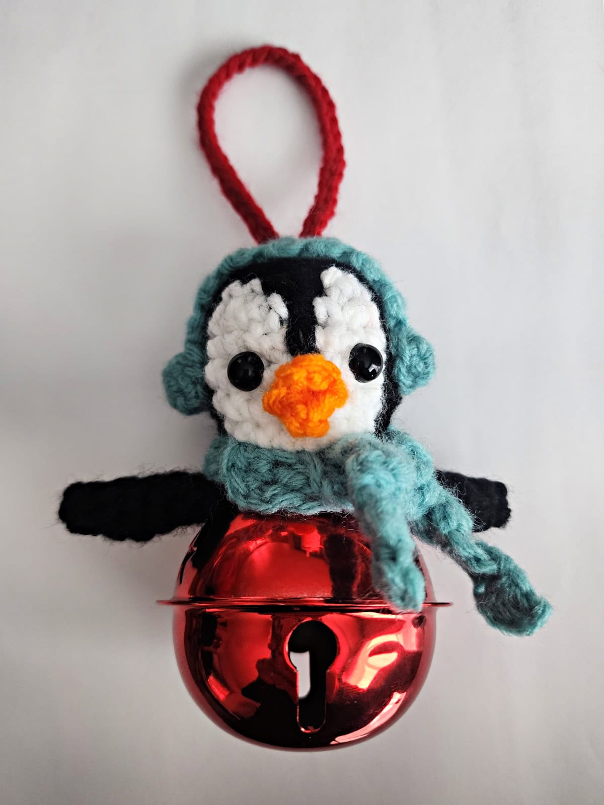 Crochet penguin jingle bell ornament with earmuffs and a scarf laying on a white table.