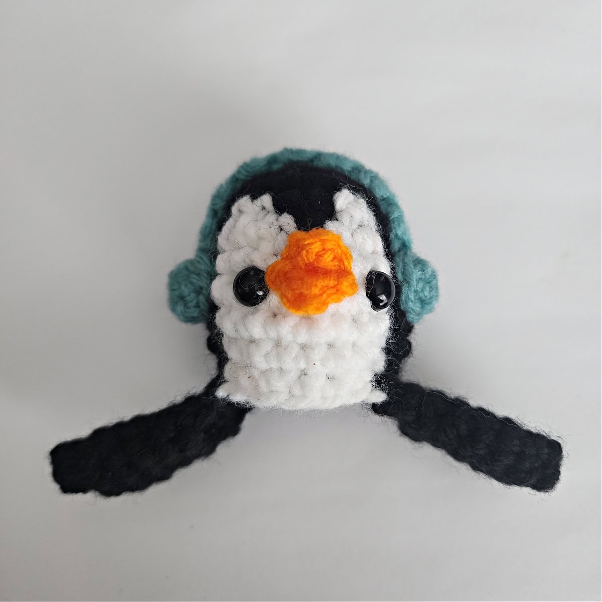 Crochet amigurumi penguin head and wings with blue earmuffs laying on a white surface.