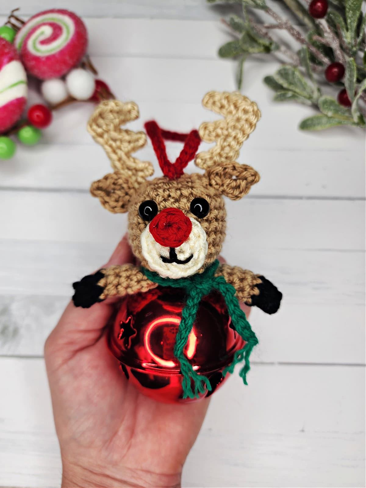 Crochet reindeer jingle bell ornament being held in hand with Christmasy background.