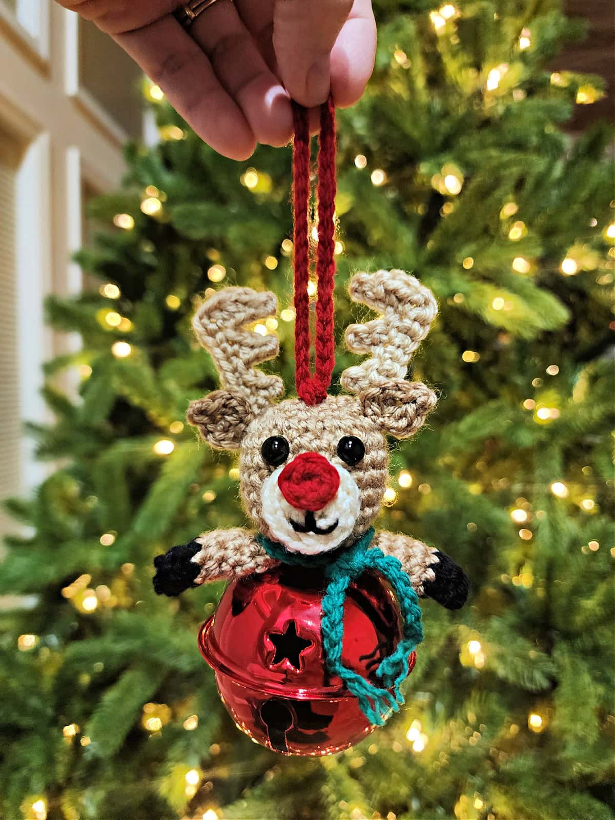 Crochet reindeer jingle bell ornament held in front of a Christmas tree.