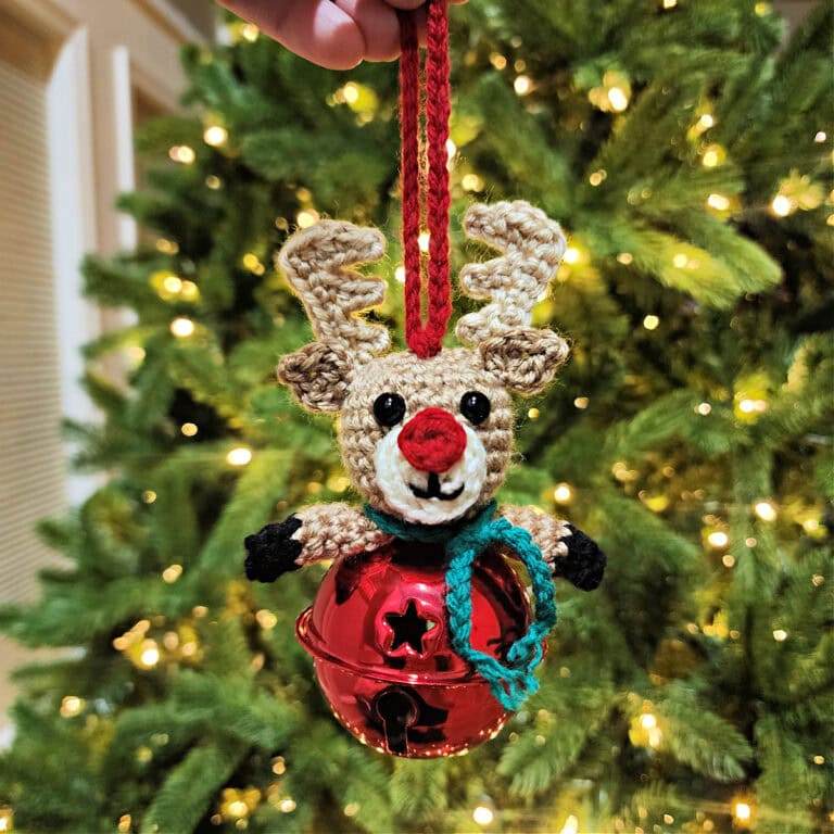 Crochet reindeer jingle bell ornament held in front of a lit Christmas tree.