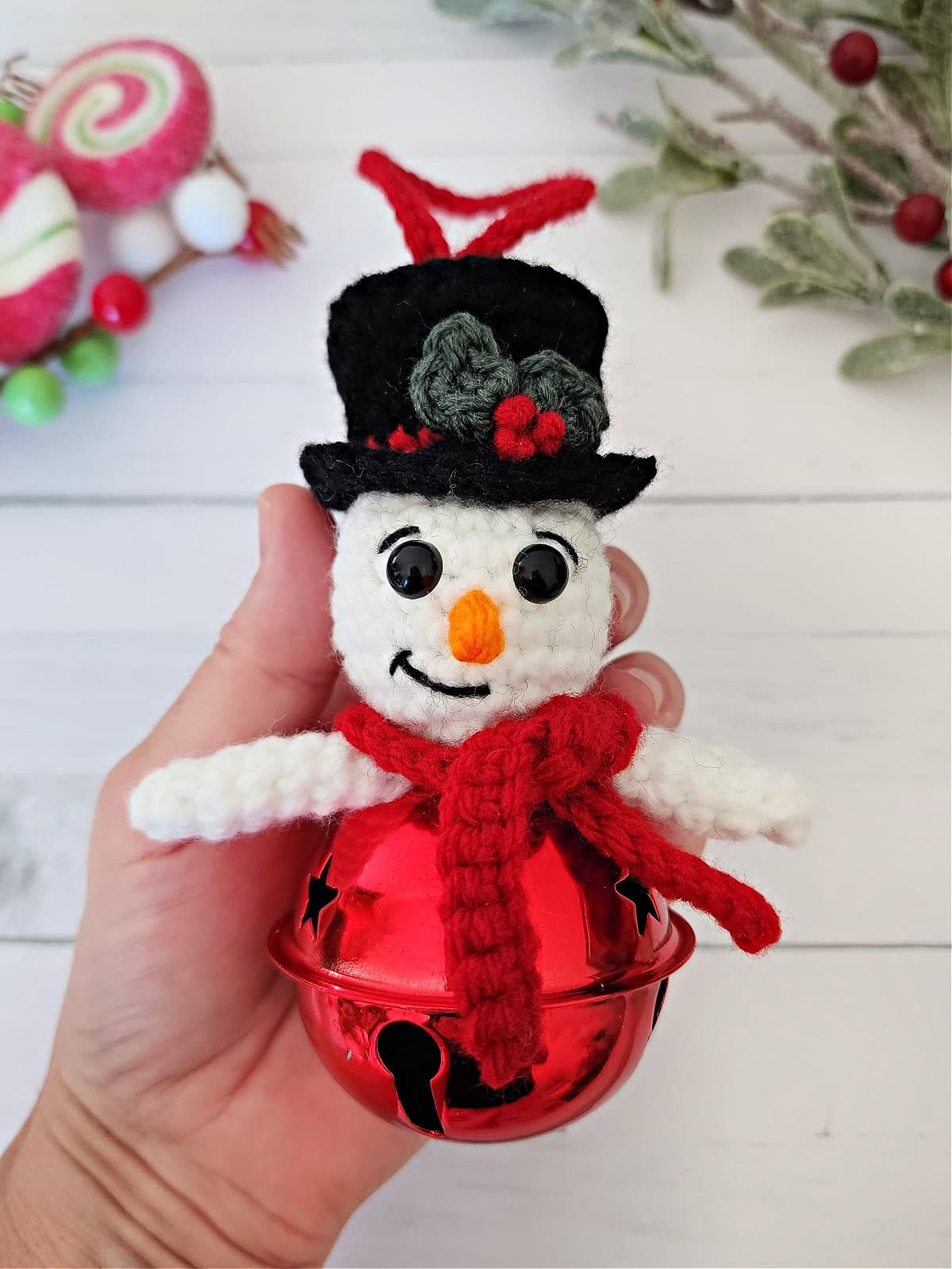 Crochet snowman jingle bell ornament being held in a hand by Christmas decor.