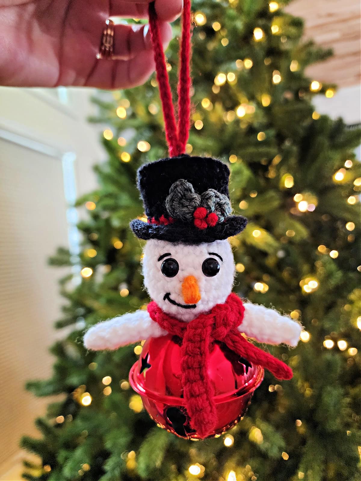 Crochet snowman jingle bell ornament being held out in front of a Christmas tree.