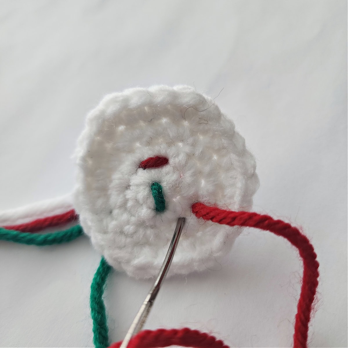 Small white crochet circle and yarn needle adding red and green sprinkles in the icing hair.