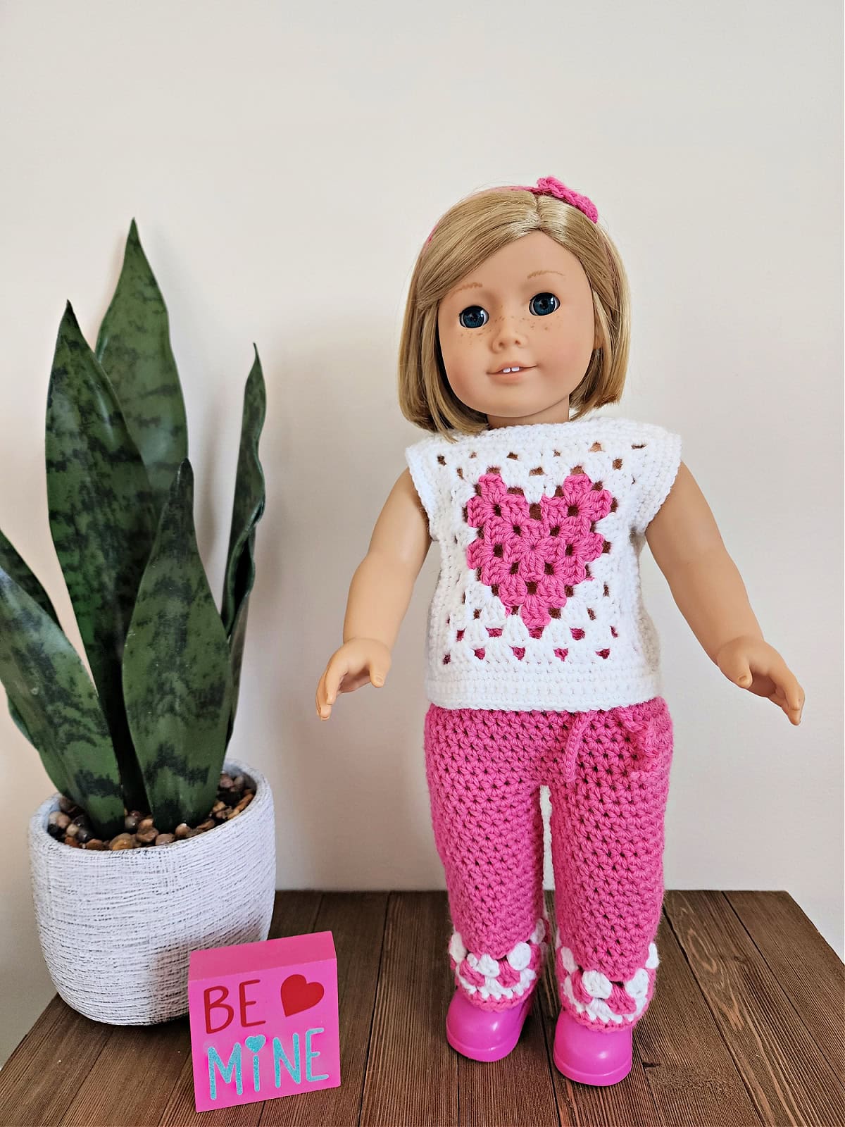 18" doll wearing crochet doll clothes and standing by a potted plant and Valentine's sign.