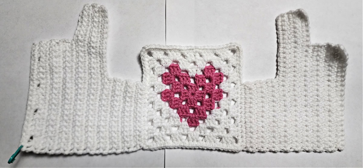 White crochet doll sweater with pink heart on the center, ready for shoulders to be seamed.