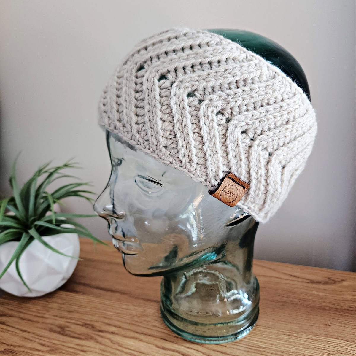 Knit-look crochet ear warmer with a leather label on a glass mannequin head.