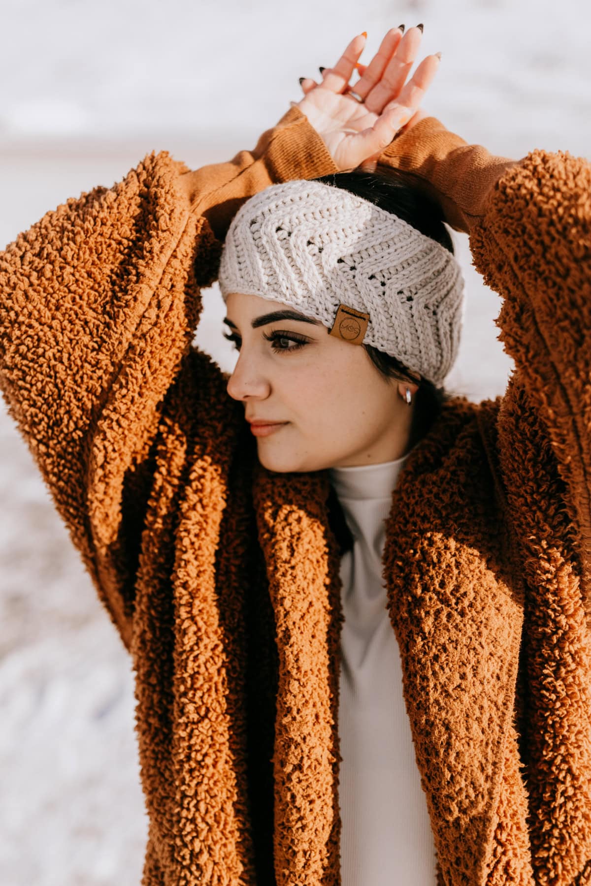 Knit-look crochet ear warmer in a linen color yarn modeled by a woman in a rust colored jacket.