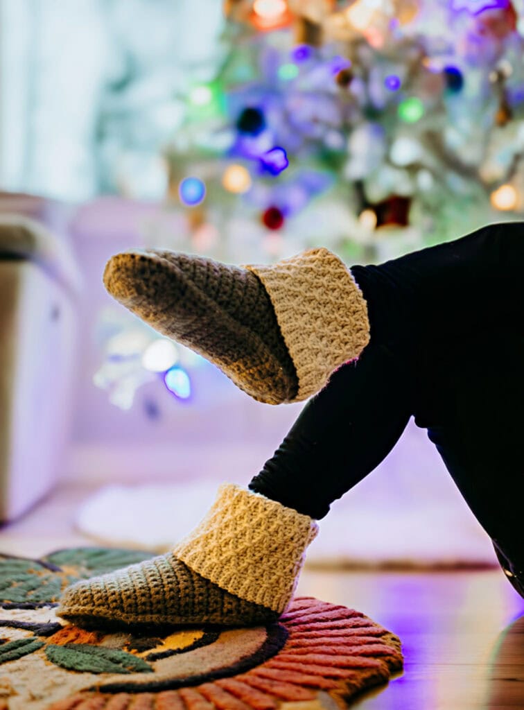 Model wearing slipper boots and leggings with leg kicked up in front of Christmas tree.