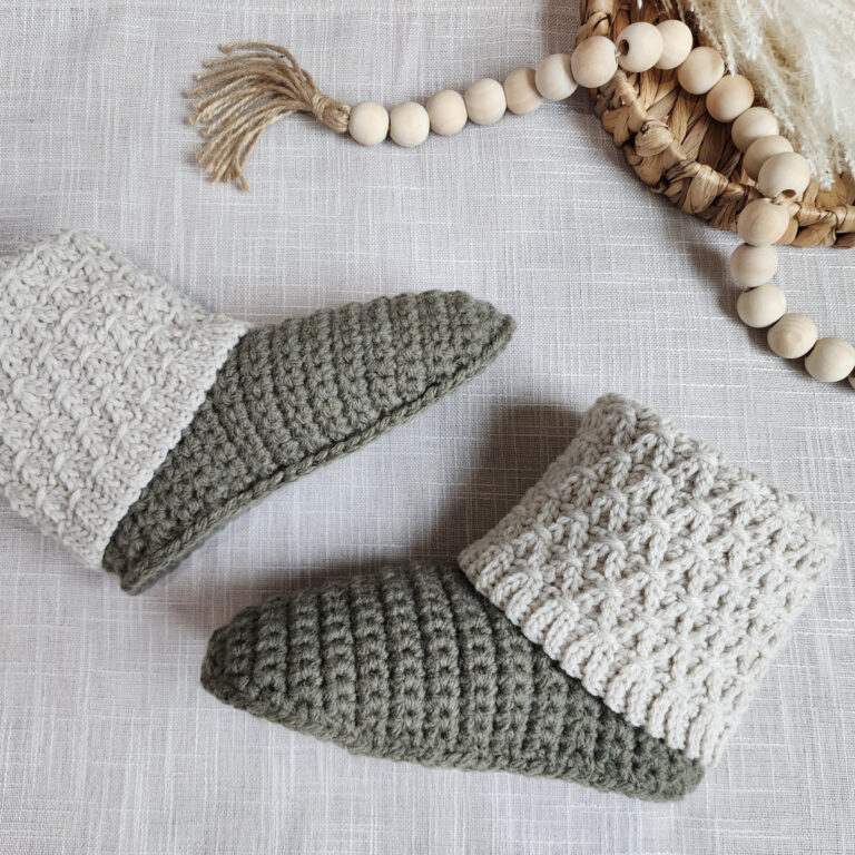 A pair of crochet slippers with knit-look cuffs laying on woven fabric next to wooden beads.