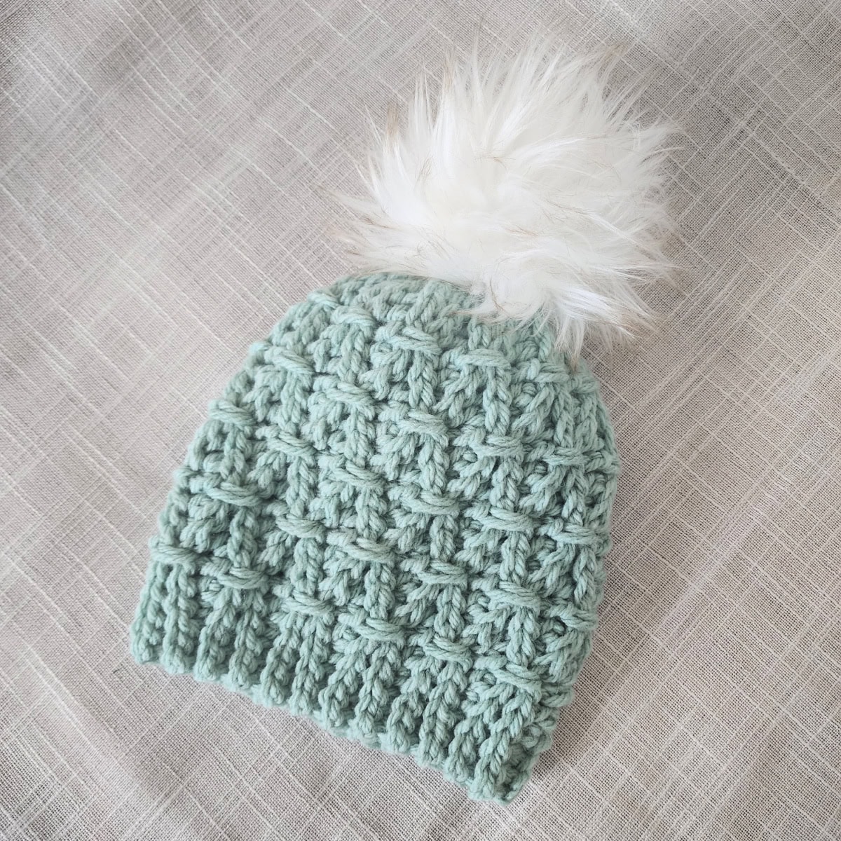 A succulent green crochet hat with a white faux fur pom laying on woven fabric.