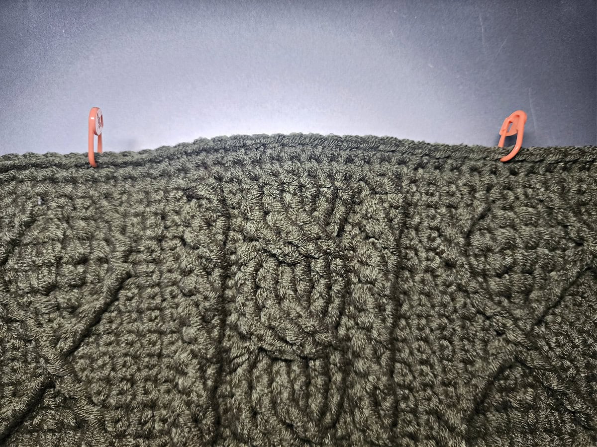 The top of a green crochet bag with two orange stitch markers marking where the yarn straps will be added.