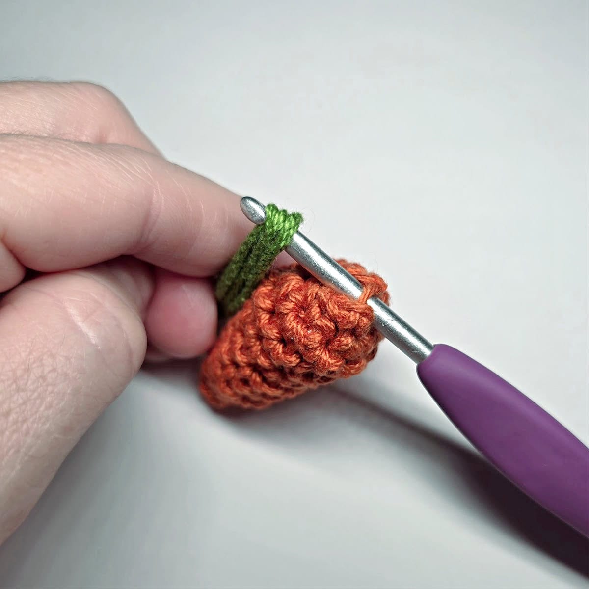 A purple crochet hook showing how to pull strands of yarn through to create the carrot top.