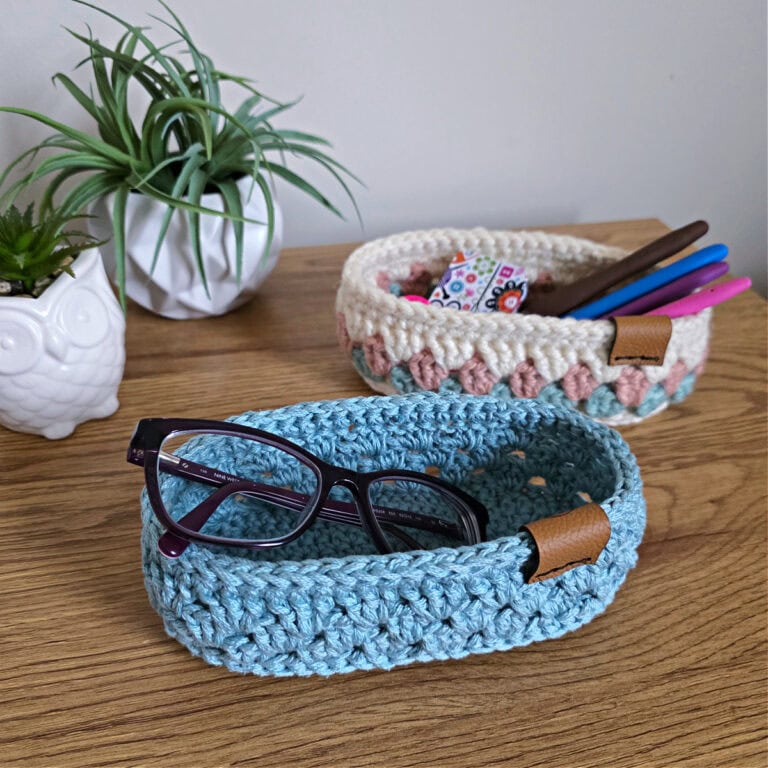 A blue crochet glasses holder and cream granny stitch glasses holder on a table.