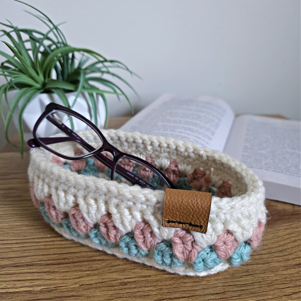 Crochet glasses holder on a table with an open book and a plant.